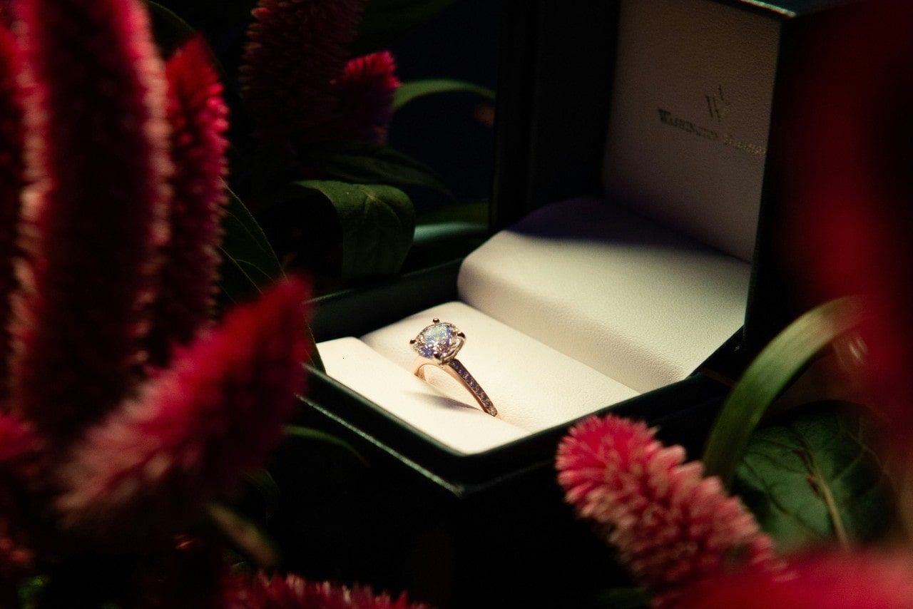 A diamond engagement ring displayed inside an open green velvet box surrounded by red flowers.