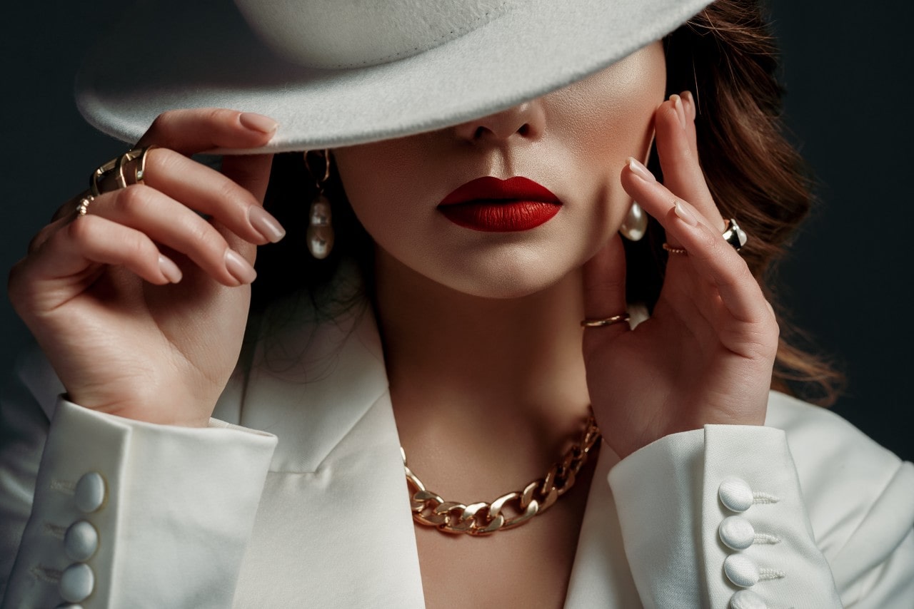 A woman in a white suit and hat partially covering her face, with bright red lips, pearl earrings, yellow gold chain necklace, and rings.