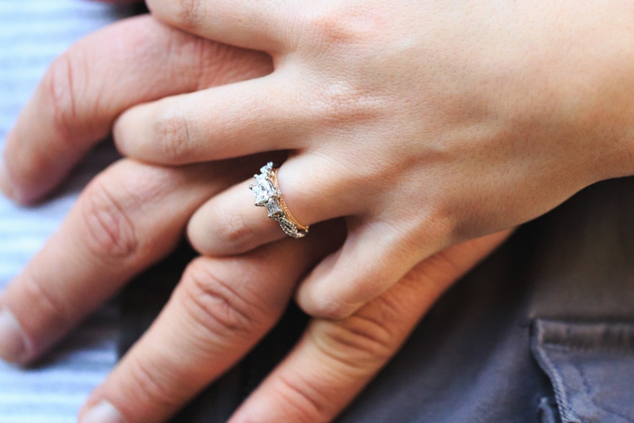 a woman’s hand over a man’s, the woman’s donning a mixed metal diamond engagement ring
