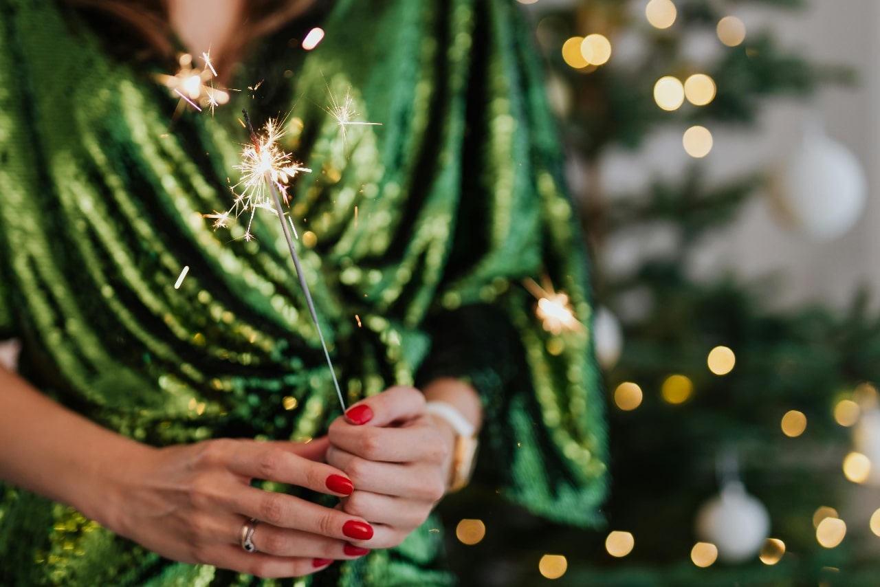 A close-up of a festively dressed woman in a green dress holding a lit sparkler among a sparkling Christmas tree.