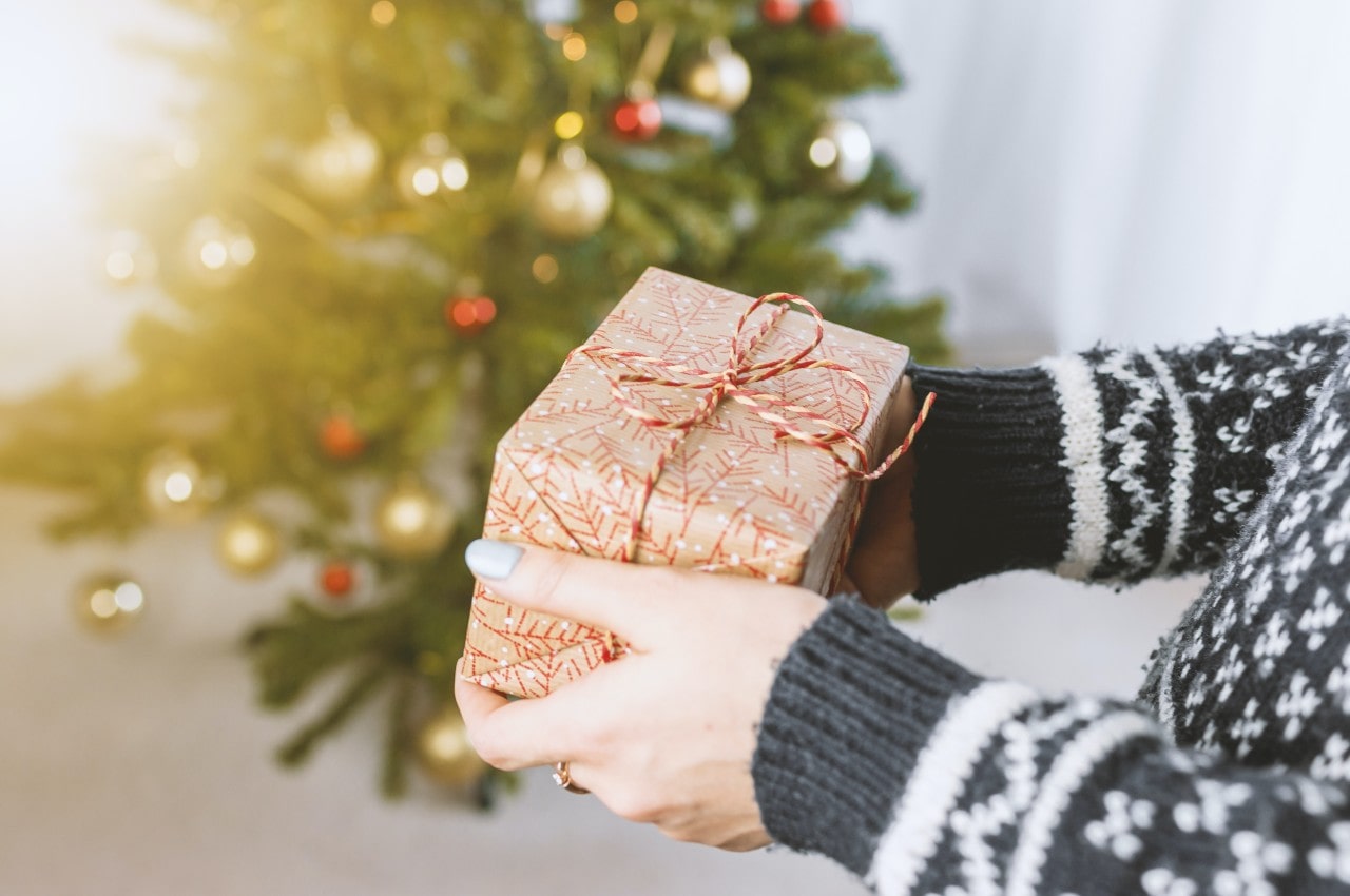 A close-up of a woman wearing a winter sweater holding a wrapped gift in front of a decorated Christmas tree.