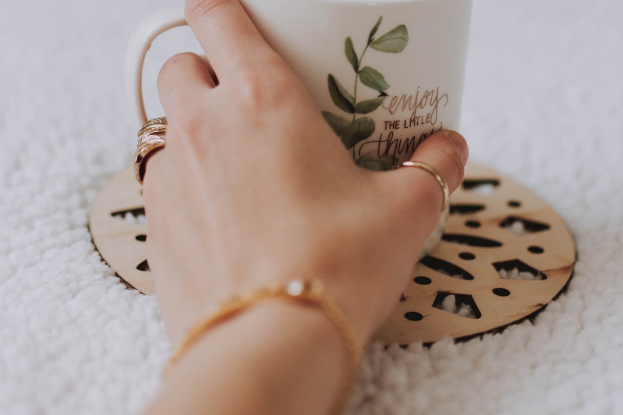 A close-up of a person’s hand, adorned with multiple rings and a delicate chain bracelet, reaching for a coffee mug.