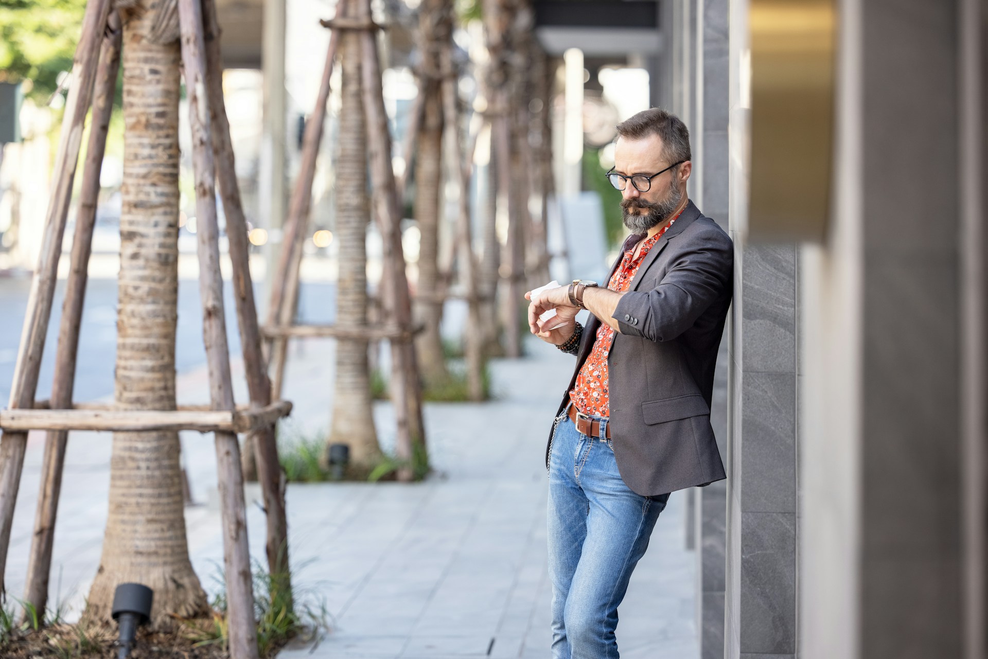 A wide angle view of a casually dressed man checking his watch as he leans on a building.