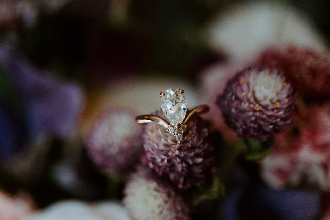 A pear-shaped diamond engagement ring with a gold band is put atop purple clover flowers in a softly blurred floral background.