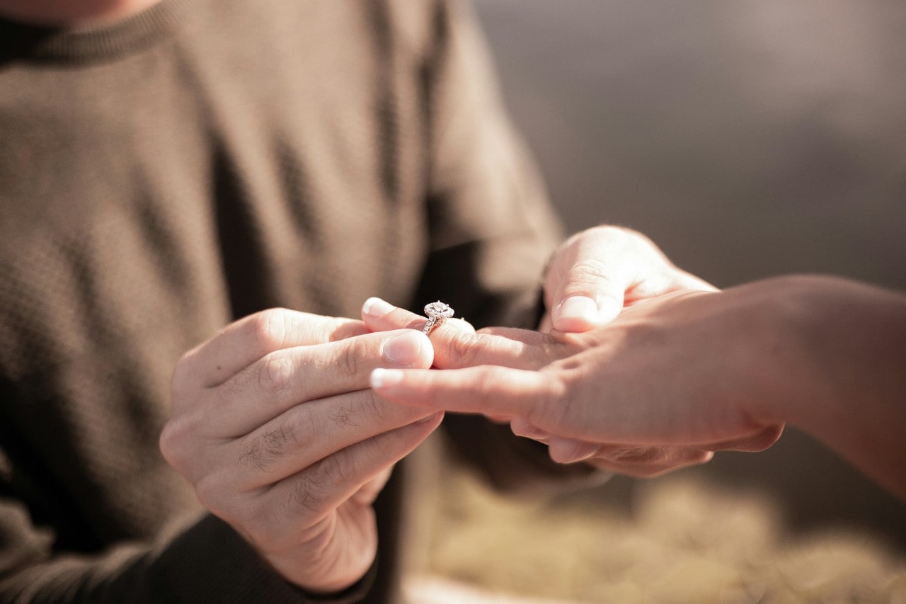 A man in a brown sweater places a halo diamond engagement ring on a woman's finger during a proposal, with natural lighting.