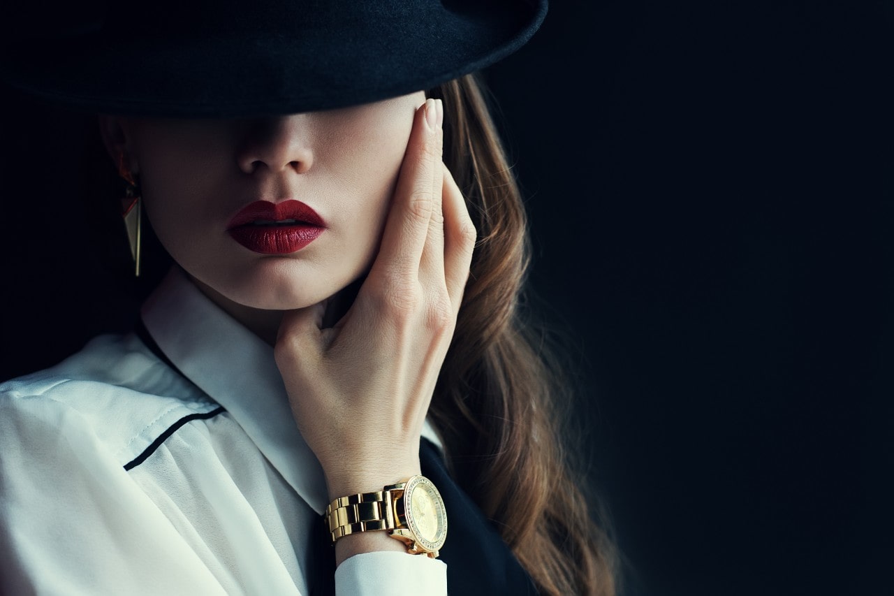 a woman in a black hat touching her face and wearing a bold yellow gold watch