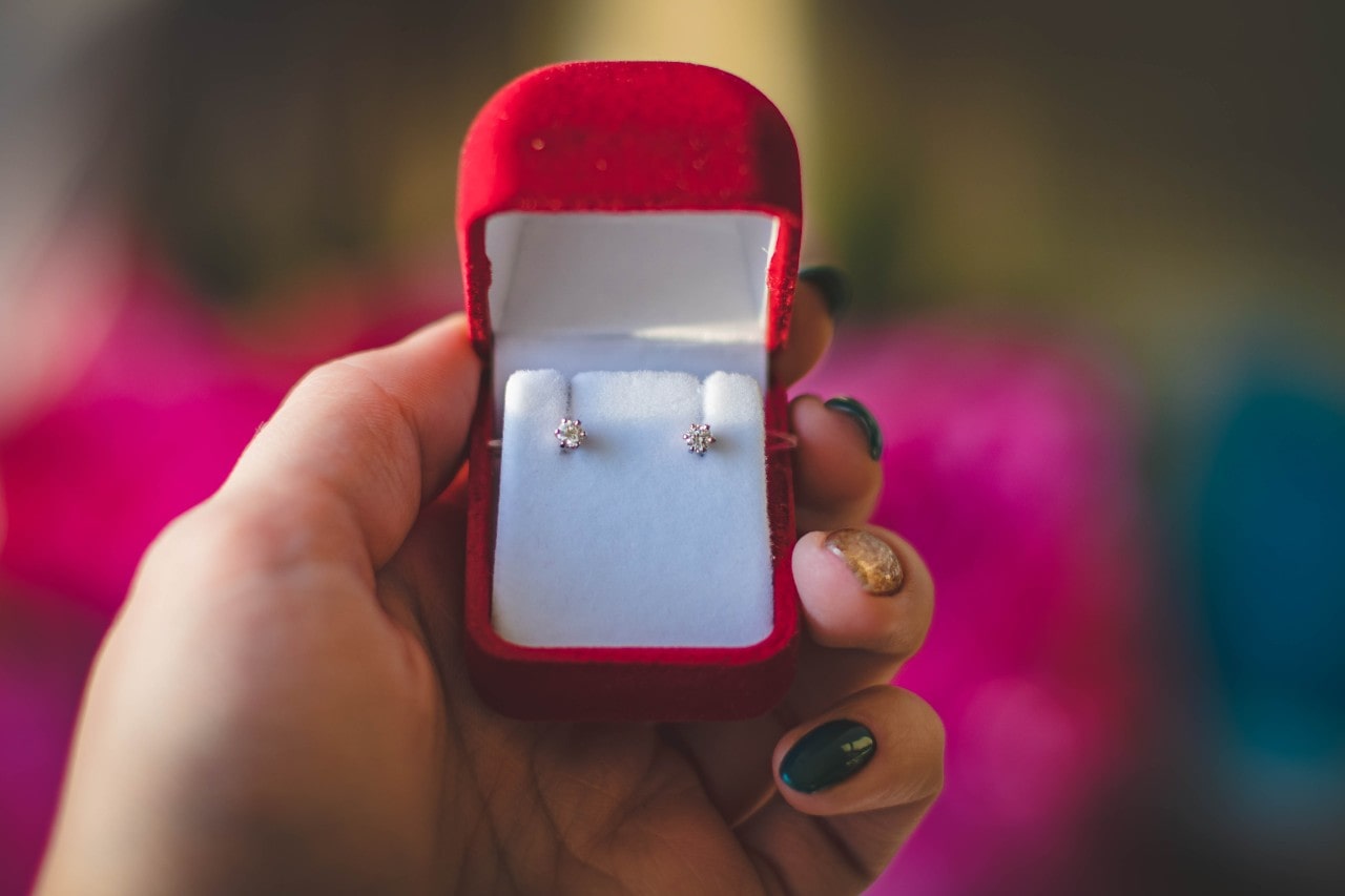 a pair of diamond stud earrings sit in a jewelry box in a woman’s hand.