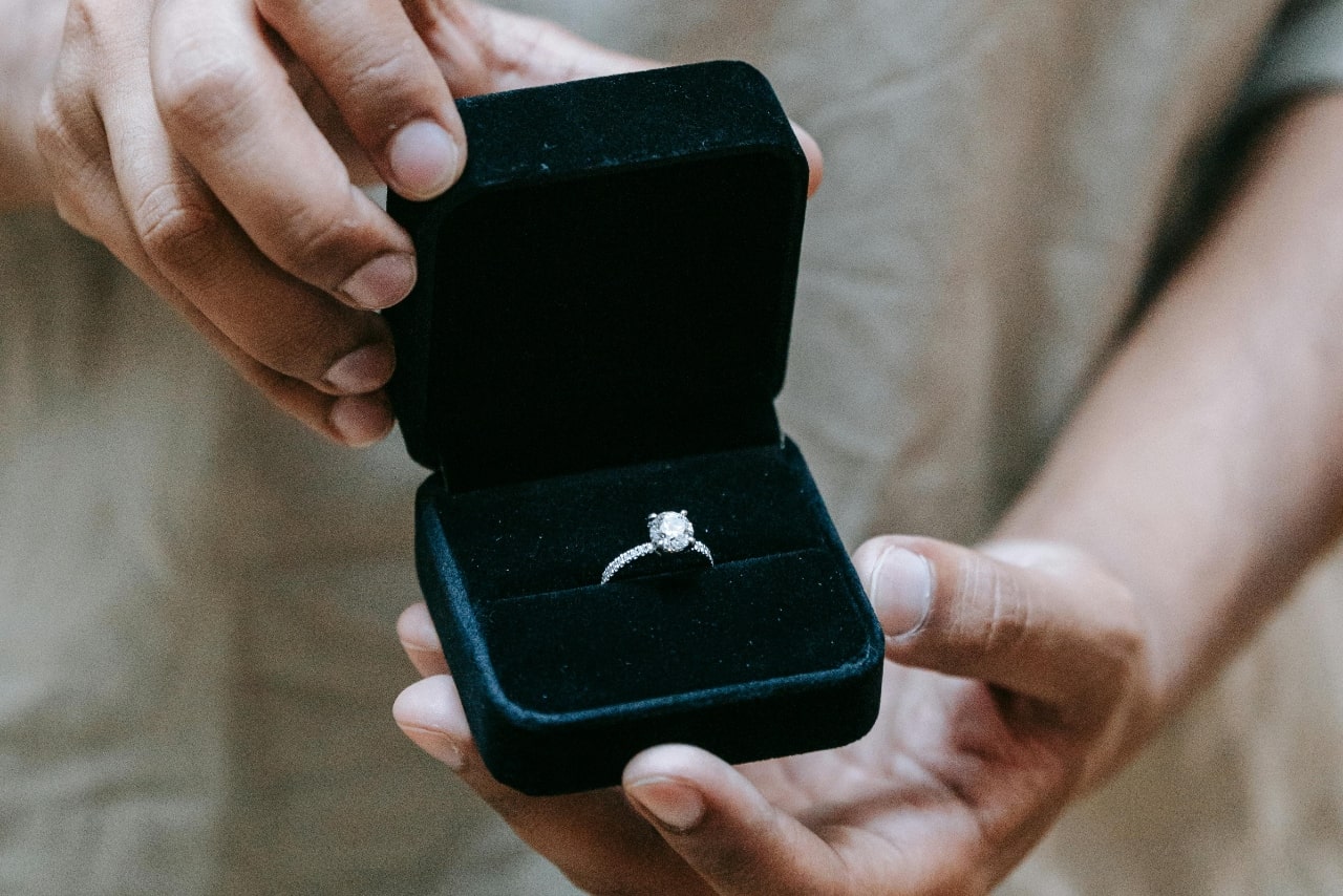A person holding an open black velvet box displaying a sparkling diamond engagement ring.