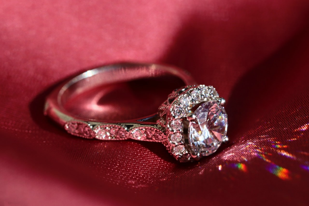 A diamond halo engagement ring with a round brilliant center stone sparkles on a deep red satin background.