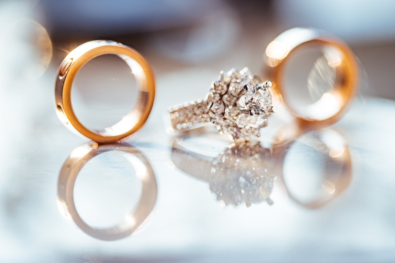 A close-up of two gold wedding bands and a diamond engagement ring placed on a reflective surface with soft light.