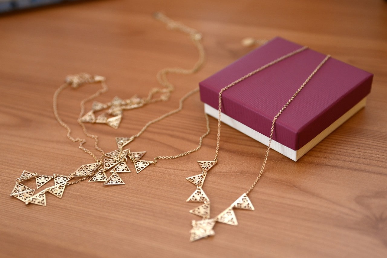 Two gold necklaces with triangular designs displayed beside a purple jewelry box on a wooden surface Two gold necklaces with triangular designs displayed beside a purple jewelry box on a wooden surface