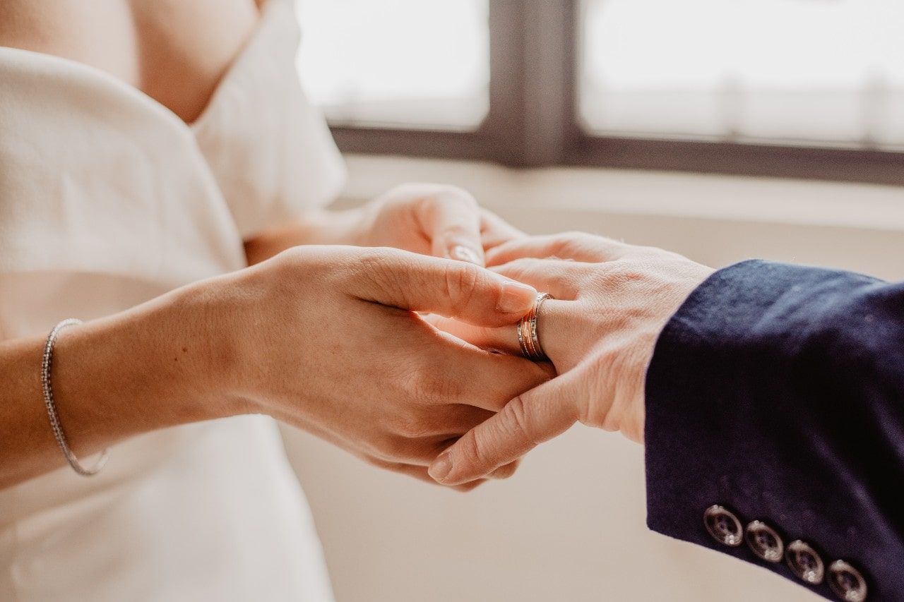 A bride places a mixed metal wedding band on the groom's finger during a ceremony, with natural light streaming through a window in the background. A bride places a mixed metal wedding band on the groom's finger during a ceremony, with natural light streaming through a window in the background.