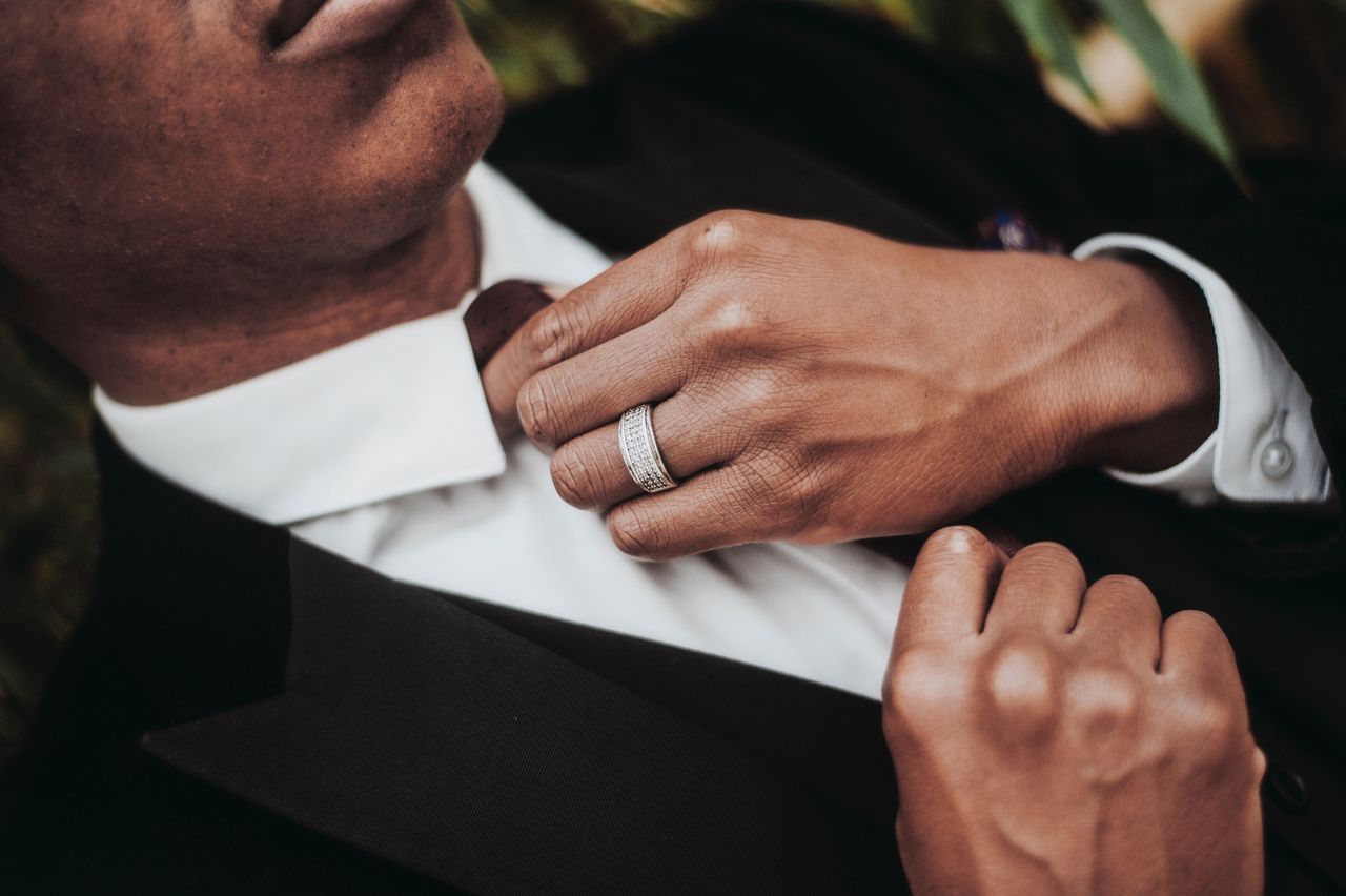 A groom in a black tuxedo adjusts his tie, showcasing a detailed white gold diamond wedding band on his finger. A groom in a black tuxedo adjusts his tie, showcasing a detailed white gold diamond wedding band on his finger.