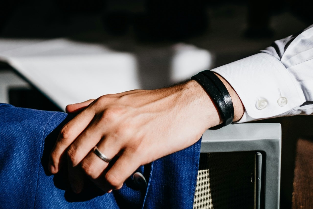 A man rests his hand on a table, showcasing a polished wedding band and black leather bracelet. A man rests his hand on a table, showcasing a polished wedding band and black leather bracelet.