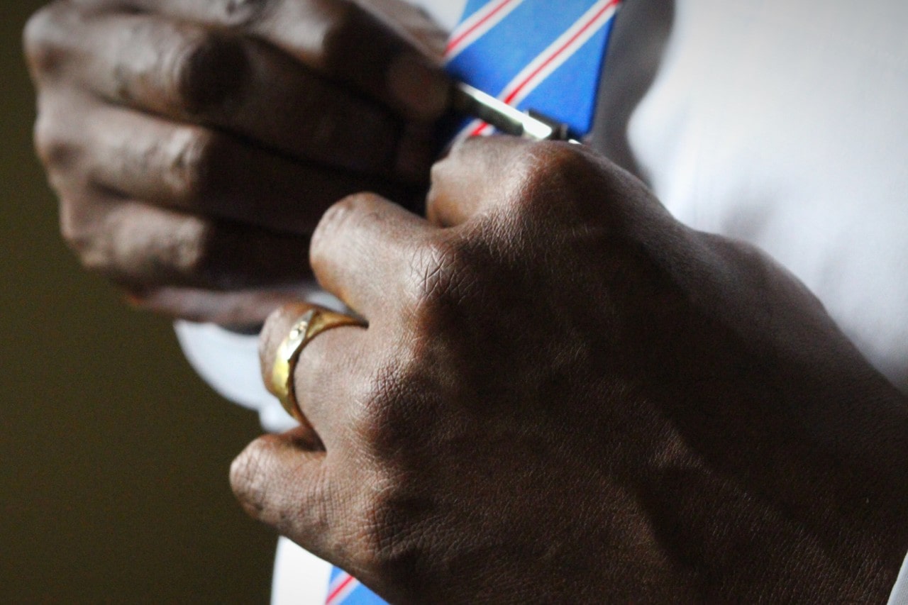 A close-up of a groom adjusting his tie clip while wearing a gold wedding band and a white dress shirt with a colorful striped tie. A close-up of a groom adjusting his tie clip while wearing a gold wedding band and a white dress shirt with a colorful striped tie.