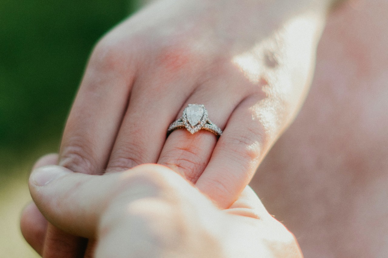 A close-up of a pear-shaped diamond engagement ring with a halo setting worn on a hand held gently in natural sunlight.