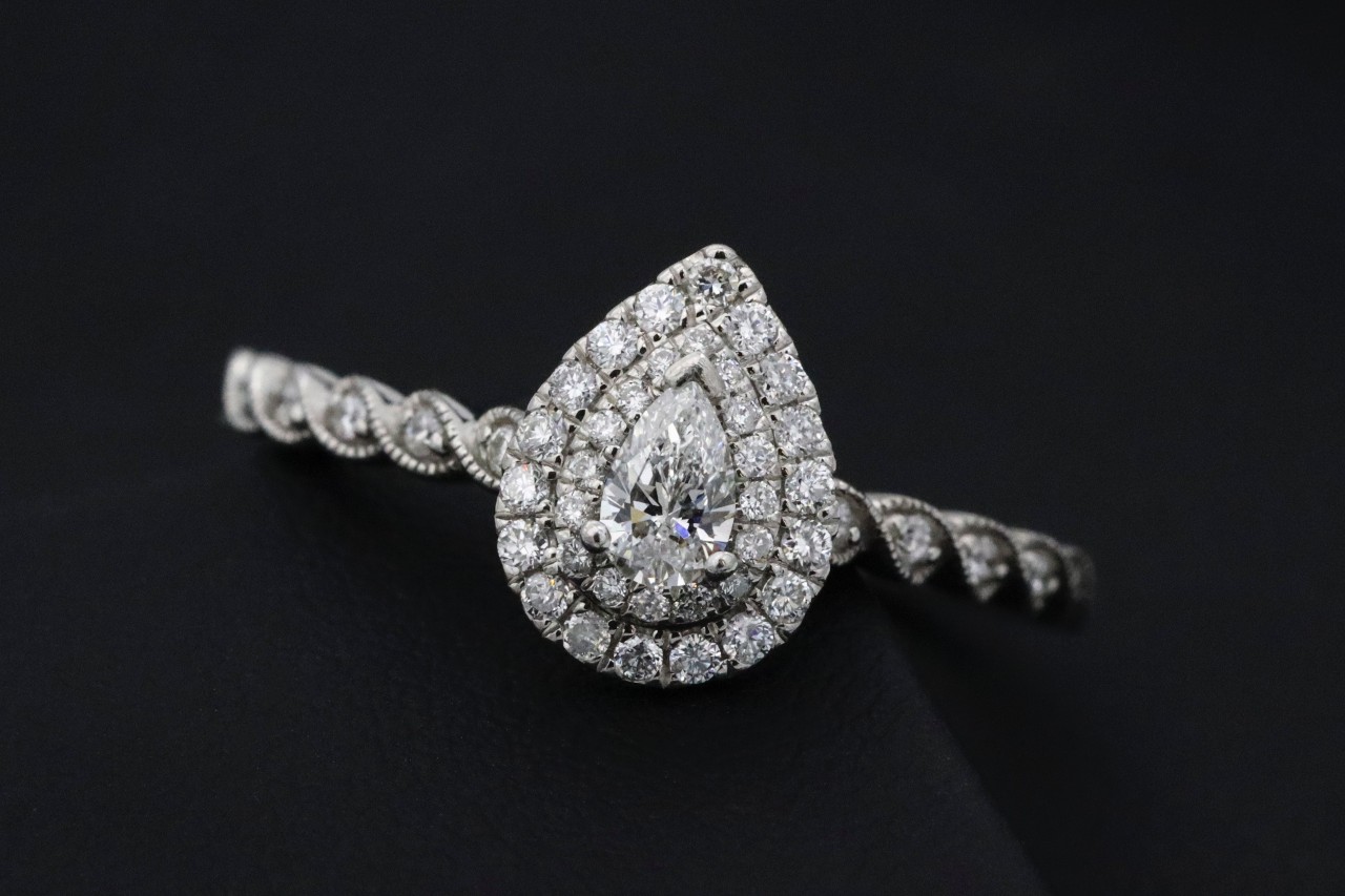 A close-up of a white gold pear-shaped diamond ring with a double halo and twisted diamond-studded band set against a black background.