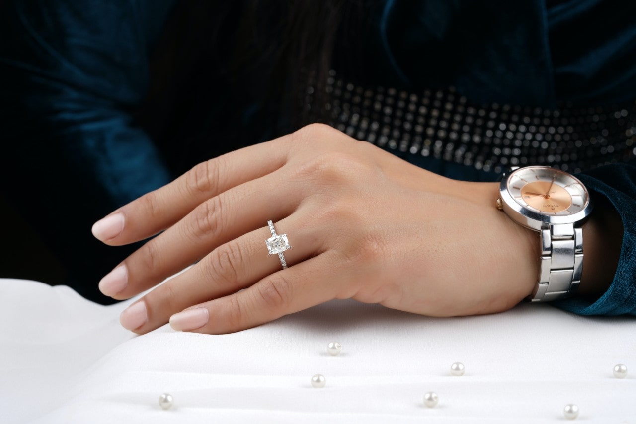 A woman's hand adorned with a radiant cut diamond engagement ring and a stainless steel wristwatch, resting gently on a white surface decorated with scattered pearls.