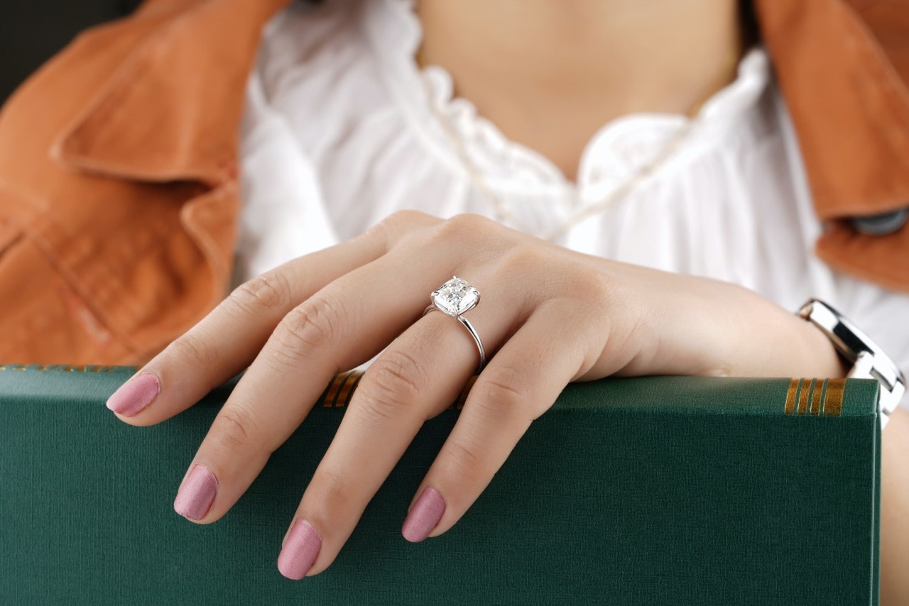 A woman’s manicured hand resting on a green book, a solitaire ring with a radiant cut center stone on her finger.