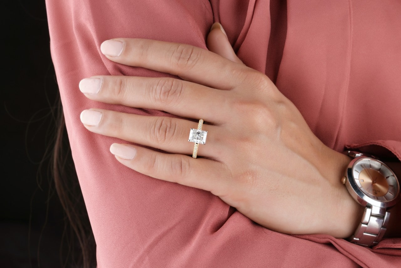 A woman's hand adorned with a yellow gold radiant cut ring resting against her pink blouse, a stainless steel watch on her wrist.