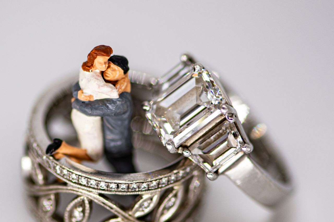 A close-up of a three stone engagement ring displayed next to two wedding bands around a small wedding figurine. A close-up of a three stone engagement ring displayed next to two wedding bands around a small wedding figurine.